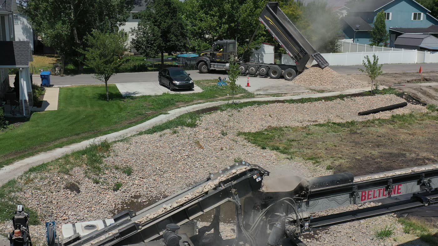 Beltline conveyor slinging gravel across a residential lot while a dump truck feeds the hopper